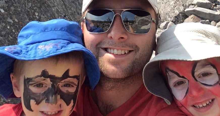 A person wearing sunglasses hugging two children with face paint and hats, smiling together outdoors.
